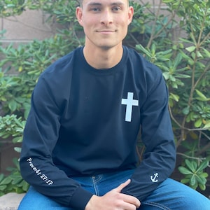 May include: A black long-sleeve shirt with a white cross on the front and white text on the sleeves. The text on the left sleeve reads "Proverbs 27:17" and the text on the right sleeve is an anchor symbol.