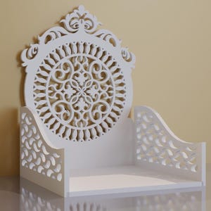 May include: White decorative shelf with intricate cut-out designs. The shelf features a central circular pattern and curved side supports. The design is symmetrical and the shelf is made of a solid white material. The background is a light yellow color.