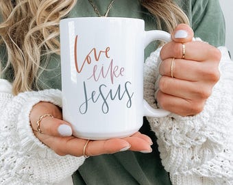 Op de afbeelding: Een witte keramische mok met een zwarte, roze en bruine inscriptie die "Love like Jesus" luidt.