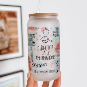 Op de afbeelding: Een matglazen beker met een bamboe deksel en een doorzichtig rietje. De beker heeft de tekst "Director Daily Affirmations" met verschillende affirmaties en decoratieve illustraties in pastelkleuren. De beker wordt door een persoon vastgehouden.