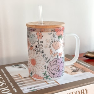 May include: A white ceramic mug with a floral pattern in shades of pink, purple, and green. The mug has a bamboo lid and a clear glass straw. The mug is sitting on a book titled "House Story".