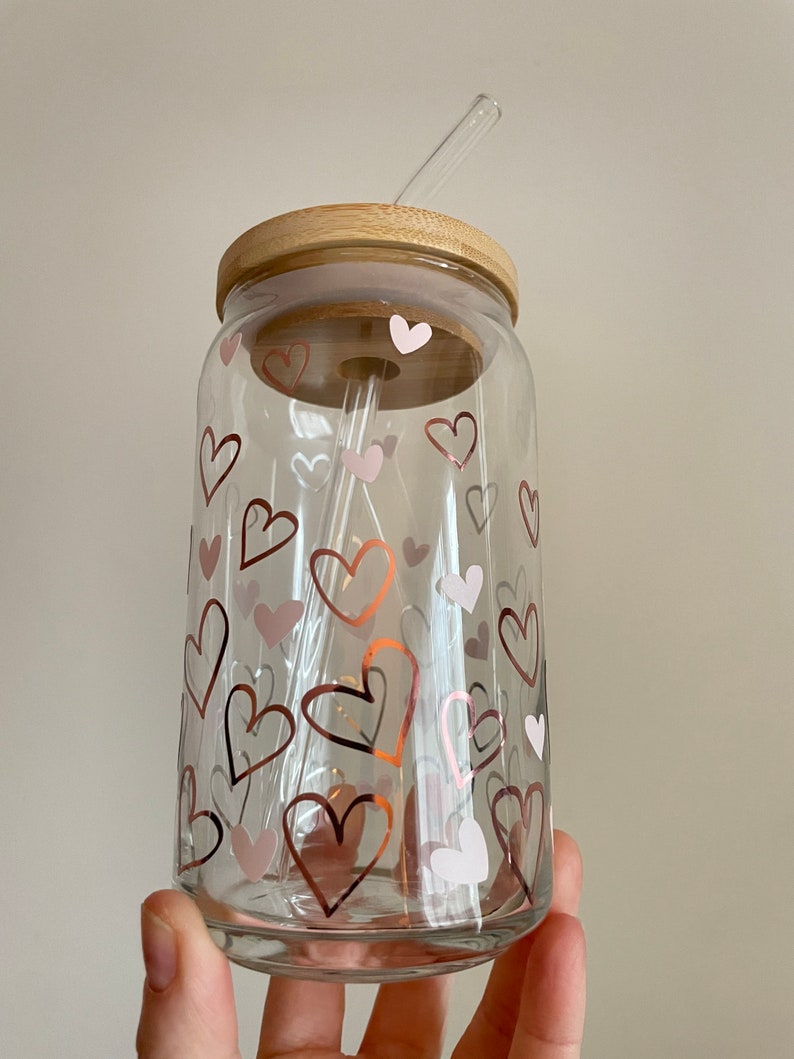 May include: A clear glass jar with a wooden lid and a straw. The jar is decorated with pink and gold hearts.