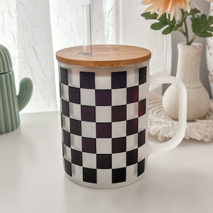 Puede incluir: Una taza de cerámica blanca con un patrón de cuadros negros y blancos. La taza tiene una tapa de bambú y una pajita de plástico transparente.