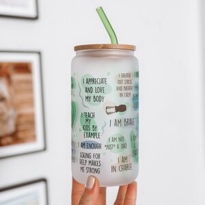 May include: A frosted glass water bottle with a bamboo lid and a green straw. The bottle has inspirational text printed on it, including "I appreciate and love my body", "I teach my kids by example", "I am brave", "I am enough", "I am not "just" a dad", "I am a leader", "Asking for help makes me strong", and "I am in charge".