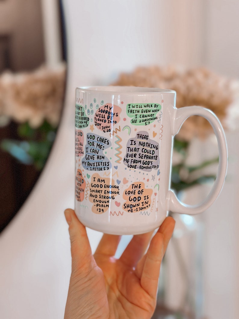 May include: White ceramic mug with colorful text and illustrations. The text includes bible verses and inspirational quotes. The mug has a handle on the right side.