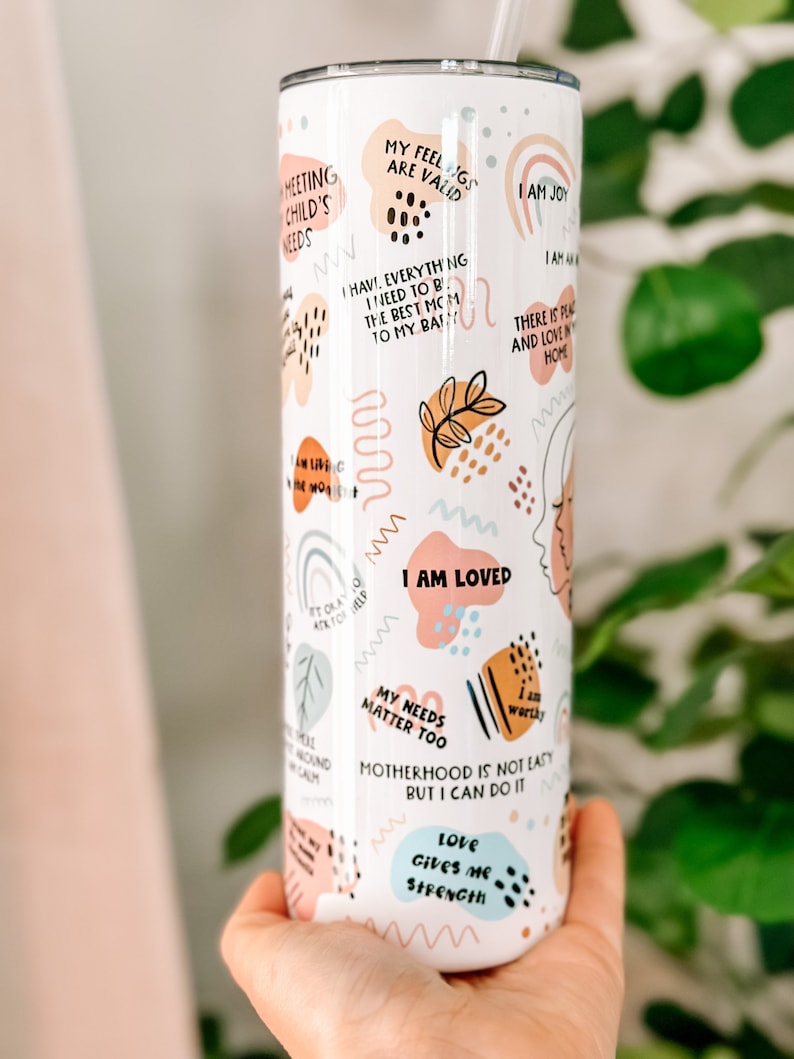 May include: White stainless steel tumbler with a colorful abstract design and motivational quotes. The quotes include "My feelings are valid", "I am loved", "Motherhood is not easy but I can do it", and "Love gives me strength".