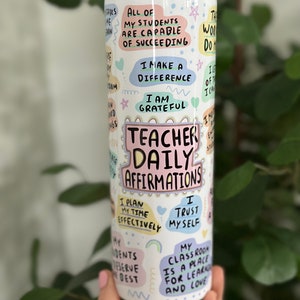 May include: White tumbler with a clear lid and a straw. The tumbler has colorful text and illustrations with positive affirmations for teachers, such as "All of my students are capable of succeeding", "I make a difference", "I am grateful", "Teacher Daily Affirmations", "I plan my time effectively", "I trust myself", and "My classroom is a place for learning and love".
