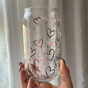 May include: A clear glass can with a pattern of pink and copper hearts. The can is being held by a hand.