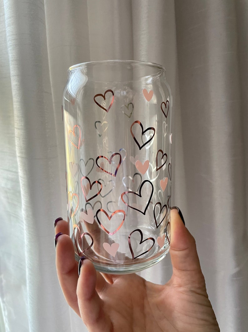 May include: A clear glass can with a pattern of pink and copper hearts. The can is being held by a hand.