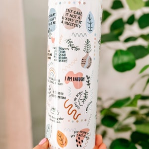 May include: White stainless steel tumbler with a variety of colorful and abstract designs and positive affirmations. The tumbler features phrases like "Self-care is not a luxury it is a necessity", "I am enough", "I love my body", and "My home is my safe place".