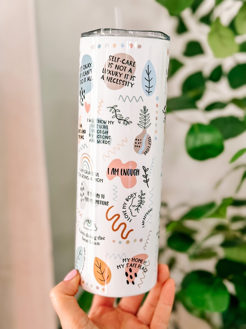 May include: White stainless steel tumbler with a variety of colorful and abstract designs and positive affirmations. The tumbler features phrases like "Self-care is not a luxury it is a necessity", "I am enough", "I love my body", and "My home is my safe place".