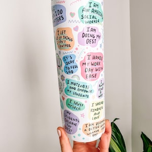 May include: A white tumbler with a straw, covered in colorful text bubbles. The bubbles contain phrases like "I am an amazing social worker" and "I motivate and empower my students." The tumbler is held by a hand.
