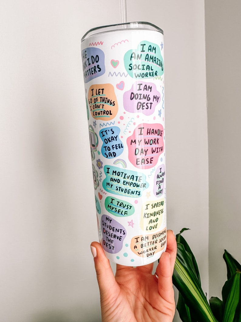 May include: A white tumbler with a straw, covered in colorful text bubbles. The bubbles contain phrases like "I am an amazing social worker" and "I motivate and empower my students." The tumbler is held by a hand.