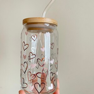 May include: A clear glass drinking cup with a bamboo lid and a glass straw. The cup has a pattern of pink and gold hearts.