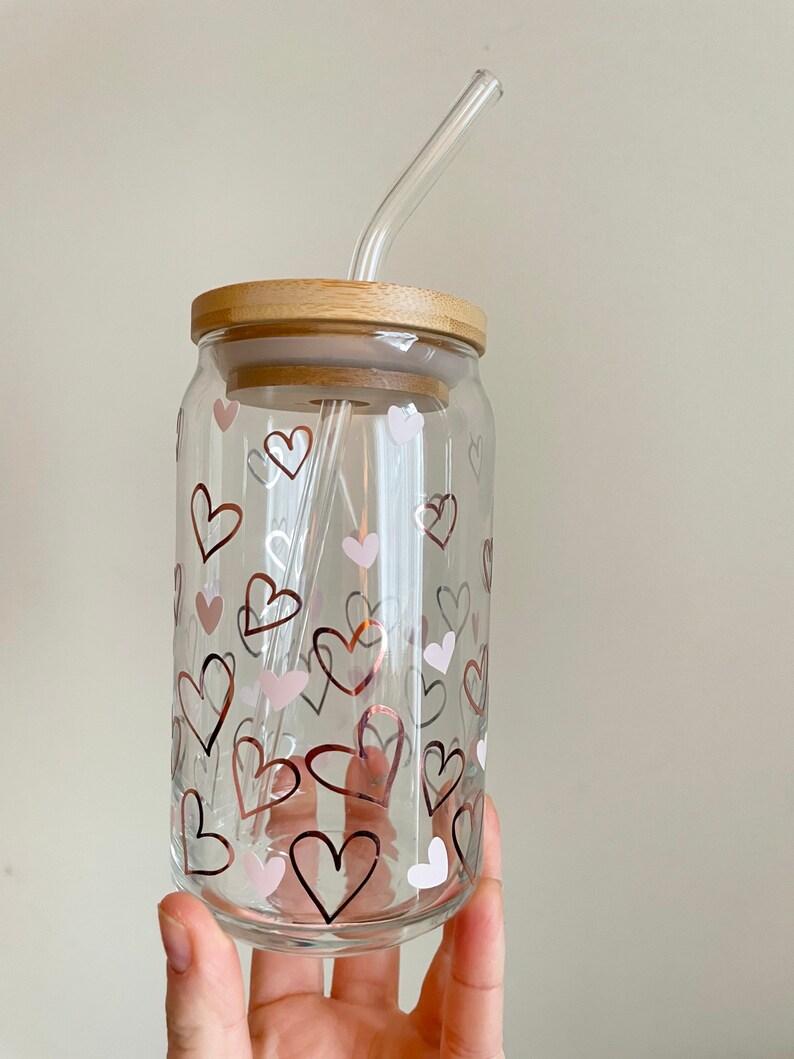 May include: A clear glass drinking cup with a bamboo lid and a glass straw. The cup has a pattern of pink and gold hearts.
