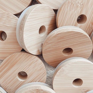 May include: A close-up of a pile of round, wooden lids with a white silicone ring around the edge. The lids are made of natural bamboo and have a hole in the center.