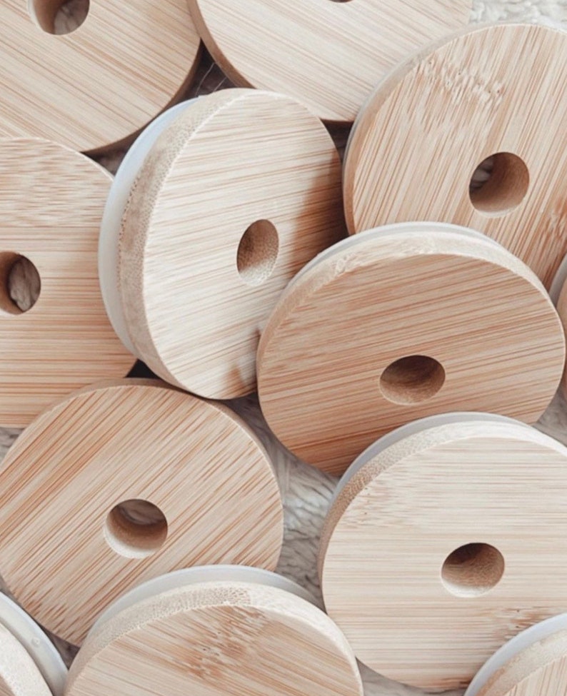 May include: A close-up of a pile of round, wooden lids with a white silicone ring around the edge. The lids are made of natural bamboo and have a hole in the center.