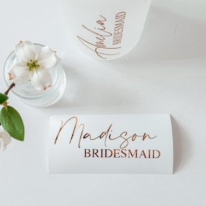 May include: A frosted glass tumbler with a bamboo lid featuring the name Amelia and the word BRIDESMAID in copper lettering, alongside a white rectangular decal with the name Madison and the word BRIDESMAID in matching copper script and print.