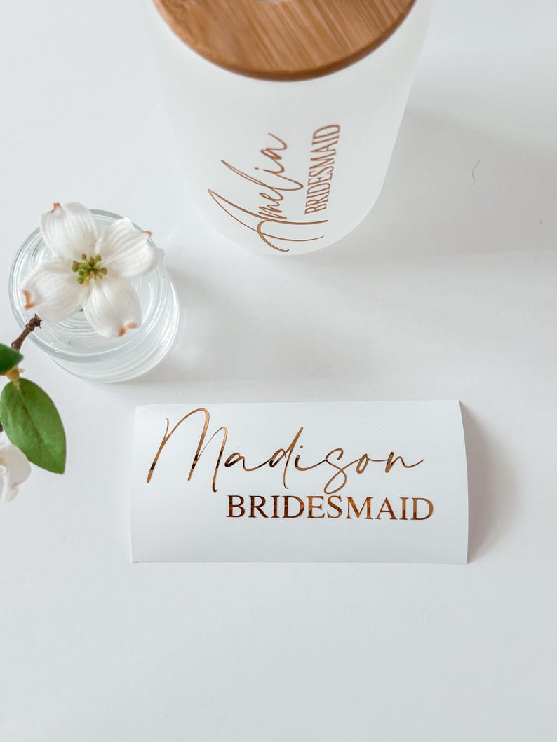 May include: A frosted glass tumbler with a bamboo lid featuring the name Amelia and the word BRIDESMAID in copper lettering, alongside a white rectangular decal with the name Madison and the word BRIDESMAID in matching copper script and print.