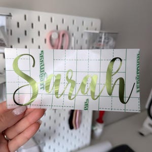 May include: A rectangular adhesive decal featuring the name Sarah in a metallic lime green cursive script font. The decal is applied to a white transfer paper backing that is printed with a green grid pattern for alignment and positioning.