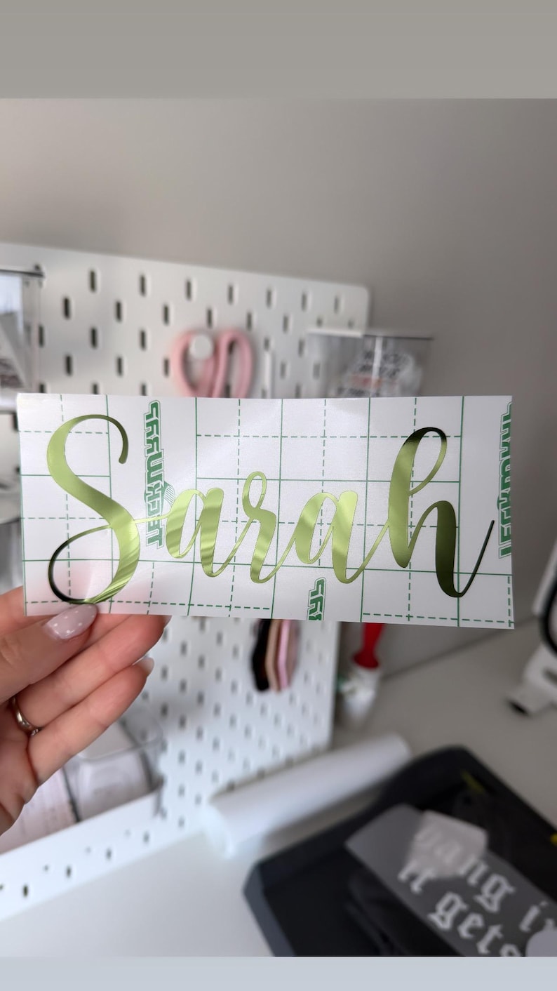 May include: A rectangular adhesive decal featuring the name Sarah in a metallic lime green cursive script font. The decal is applied to a white transfer paper backing that is printed with a green grid pattern for alignment and positioning.