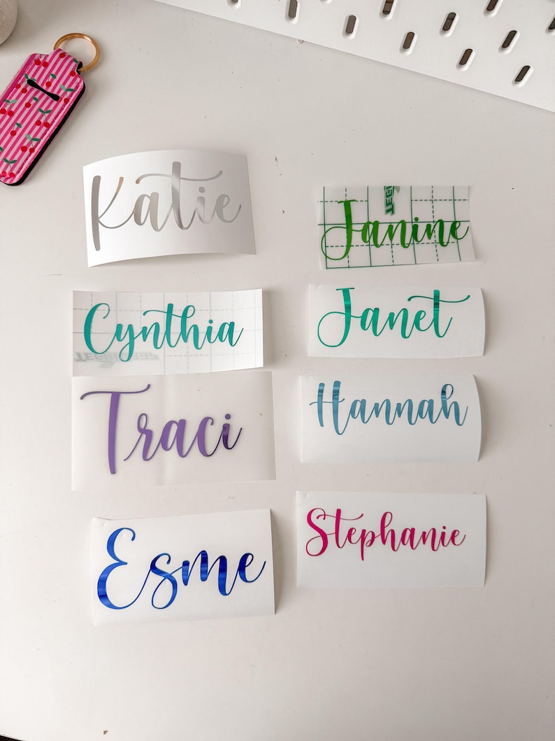 May include: A collection of eight custom vinyl name decals in various colours and script fonts, including the names Katie, Janine, Cynthia, Janet, Traci, Hannah, Esme, and Stephanie, arranged on a white surface with a cherry-patterned keychain.