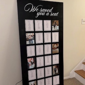 May include: A large black rectangular wedding seating chart sign with the white script text "We saved you a seat" at the top. The board features a grid of white cards displaying table numbers and guest names, interspersed with several small square photographs.
