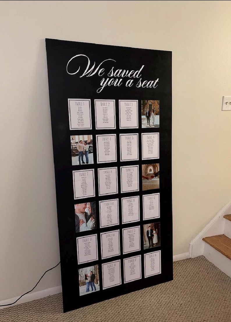 May include: A large black rectangular wedding seating chart sign with the white script text "We saved you a seat" at the top. The board features a grid of white cards displaying table numbers and guest names, interspersed with several small square photographs.