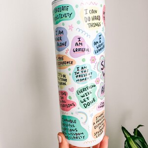 May include: A white tumbler with a silver lid and straw, covered in colorful, handwritten affirmations. Phrases include "Radiate Positivity," "I Am Grateful," and "Everything Will Get Done." The tumbler is held by a person.