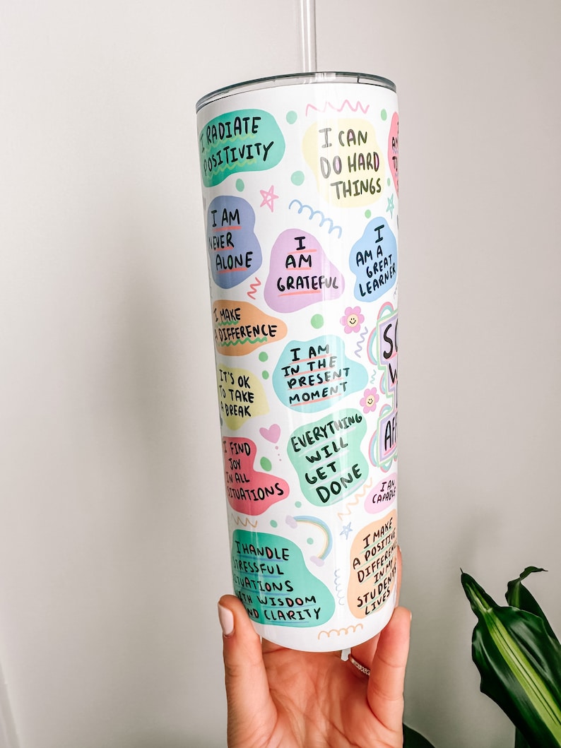 May include: A white tumbler with a silver lid and straw, covered in colorful, handwritten affirmations. Phrases include "Radiate Positivity," "I Am Grateful," and "Everything Will Get Done." The tumbler is held by a person.