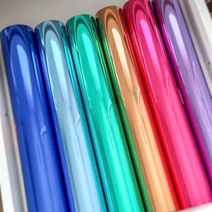 May include: A set of six cylindrical rolls of metallic foil arranged side by side. The rolls feature a smooth, reflective finish in a vibrant spectrum of colours including deep blue, light blue, emerald green, copper, bright red, and purple.