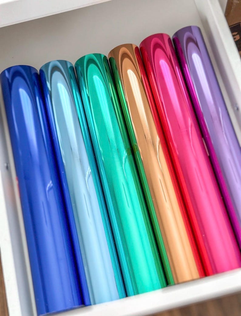 May include: A set of six cylindrical rolls of metallic foil arranged side by side. The rolls feature a smooth, reflective finish in a vibrant spectrum of colours including deep blue, light blue, emerald green, copper, bright red, and purple.