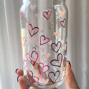 May include: A clear glass can with a pink and white heart pattern. The can is held in a hand.