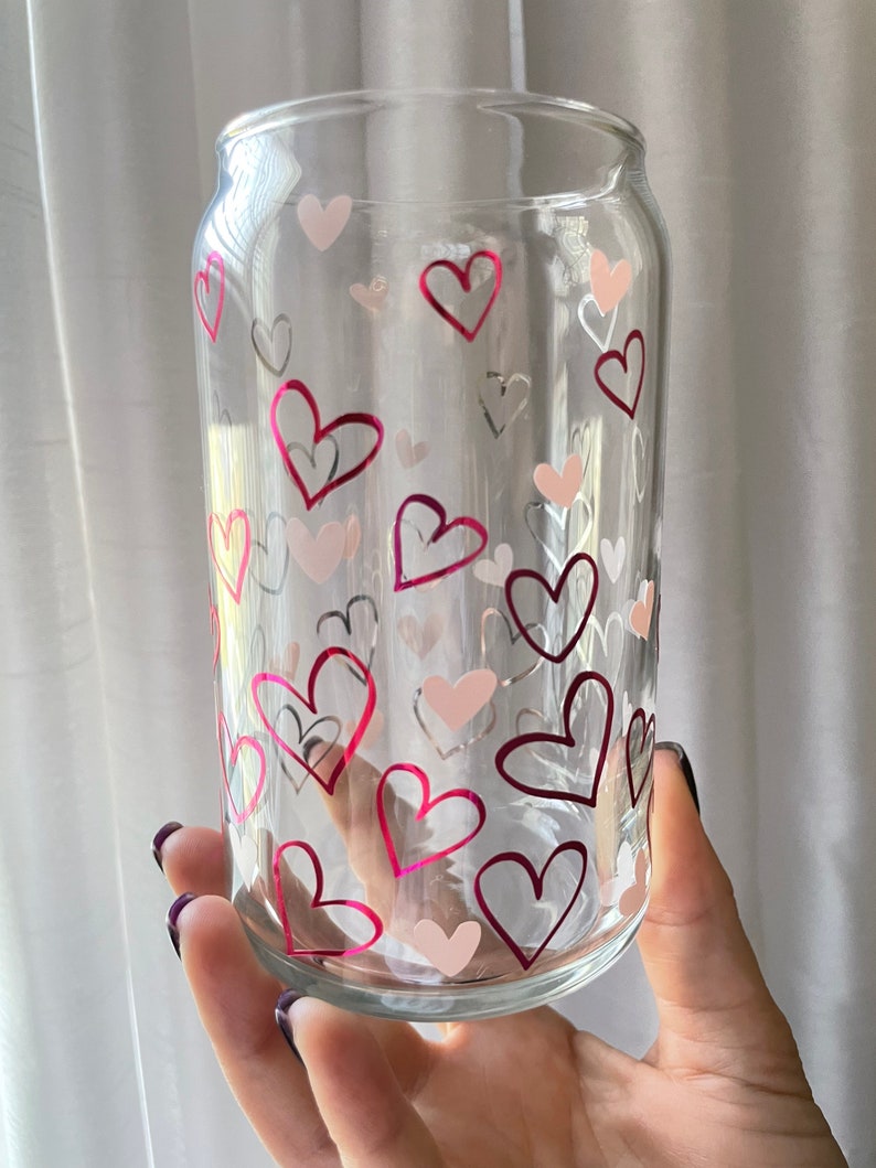 May include: A clear glass can with a pink and white heart pattern. The can is held in a hand.