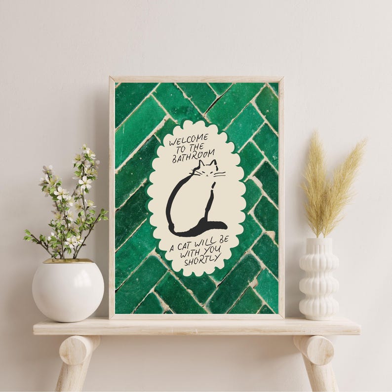 May include: A rectangular wall art print featuring a dark green herringbone tile pattern background. In the center, a cream-colored scalloped oval contains a black line drawing of a cat and the text: WELCOME TO THE BATHROOM A CAT WILL BE WITH YOU SHORTLY.