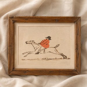 May include: A framed print depicts a rider in an orange coat and black hat on a galloping horse. The artwork is in a simple, illustrative style with brown and black lines. The frame is made of dark wood.