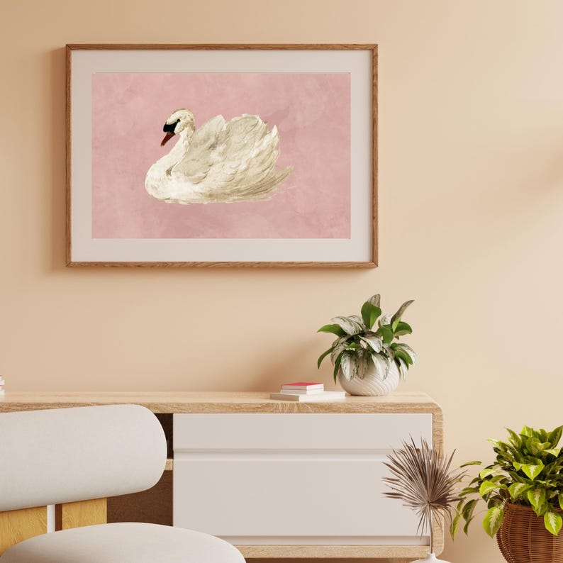 May include: A framed wall art print featuring a white swan with a black beak and eye, depicted in a resting position. The swan is set against a solid, muted pink background. The artwork is enclosed in a light-toned wooden frame with a simple border.