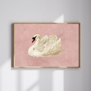 May include: A framed wall art print featuring a white swan with a black beak and orange base, depicted in a painterly style against a solid, muted dusty pink background. The artwork is enclosed in a simple, light-toned wooden rectangular frame.