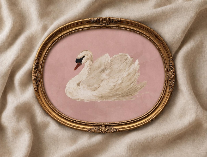 May include: An oval-shaped, ornate gold frame containing a print of a white swan with a black beak and eye detail. The swan is positioned against a solid, muted pink background, creating a contrast between the bird and the backdrop.