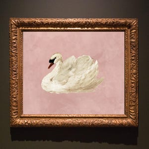 May include: A framed wall art piece featuring a white swan with a black beak and red base, set against a solid dusty pink background. The artwork is enclosed in an ornate, rectangular, metallic gold-toned frame with intricate carved detailing.