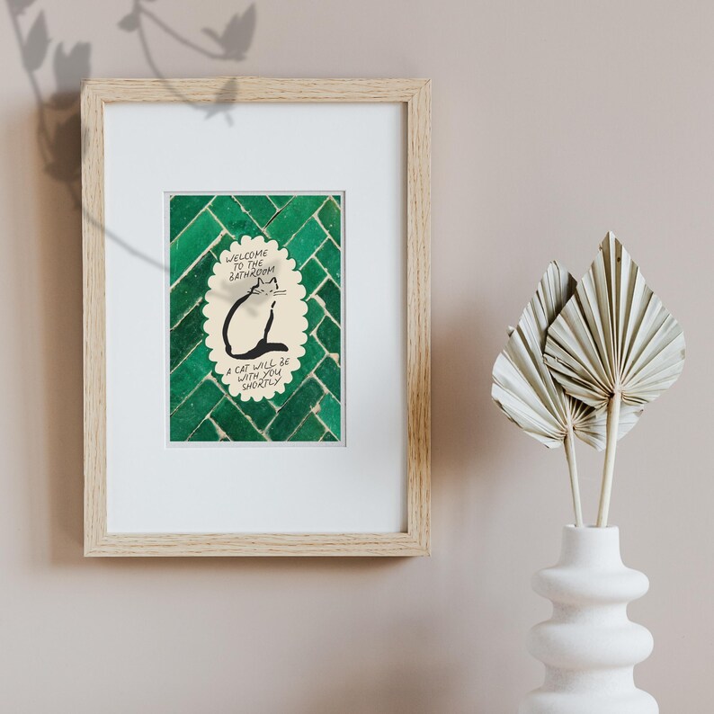May include: A framed print featuring a green tiled background and a central scalloped oval containing a black line drawing of a cat. The text reads: Welcome to the bathroom, a cat will be with you shortly. The frame is light wood with a white mat.