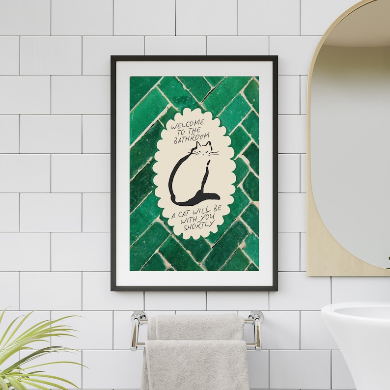 May include: A framed wall art print featuring a cream-colored scalloped oval with a black line drawing of a cat. The text reads: Welcome to the bathroom, a cat will be with you shortly. The background is a dark green, glossy, herringbone tile pattern.