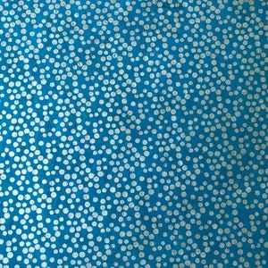 May include: A close-up of a blue fabric with a pattern of white, irregularly sized dots. The dots are scattered across the surface, creating a textured appearance. The fabric has a slightly rough texture.