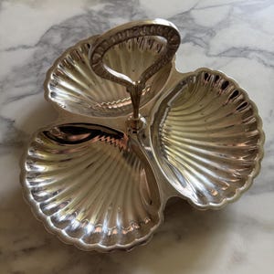 May include: Silver-plated serving dish with three scallop-shaped sections and a central handle. The dish has a reflective surface and detailed ridged patterns. The handle is arched and attached to the center.