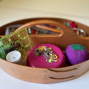 May include: A wooden sewing basket with a handle, containing sewing notions. Includes a green ruler, white thread, a pink magnetic pin holder with pins, and a purple tomato pin cushion. The basket is oval-shaped.