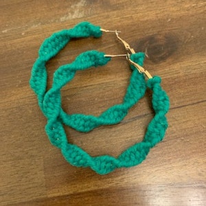 May include: A pair of green hoop earrings with a twisted rope design. The earrings are made of a soft, flexible material and have a gold-tone finish.