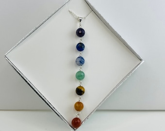 Può includere: Una collana d'argento con sette perline di pietre preziose sfaccettate multicolori. Le perline sono disposte verticalmente in un ordine arcobaleno: rosso, arancione, giallo, marrone, verde, blu e viola. La collana è presentata in una scatola bianca con bordo argentato.