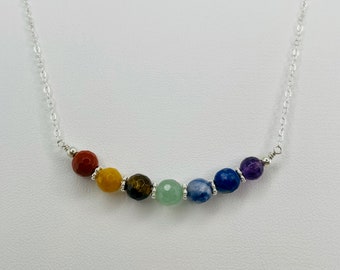 Può includere: Una collana con catena d'argento con sette perline di pietre preziose colorate. Le perline sono disposte in una linea curva, mostrando uno spettro di colori dal rosso al viola. La collana è progettata per l'uso quotidiano.