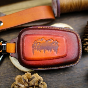 Könnte beinhalten: Ein Schlüsselanhänger aus Leder mit einem braunen Lederband und einem roten Leder-Schlüsselanhänger. Der Schlüsselanhänger hat ein Design mit einer Bergkette mit Bäumen und dem Text "4R ER".