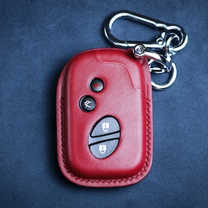 Puede incluir: Un llavero de cuero rojo con un llavero plateado y cuatro botones. Los botones tienen símbolos para bloquear, desbloquear, maletero y pánico.